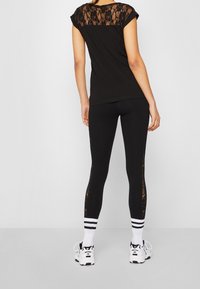 Camiseta negra con espalda de encaje, combinada con leggings negros que tienen acentos de encaje en los laterales. Calcetines de tobillo blancos a rayas y zapatillas deportivas visibles.