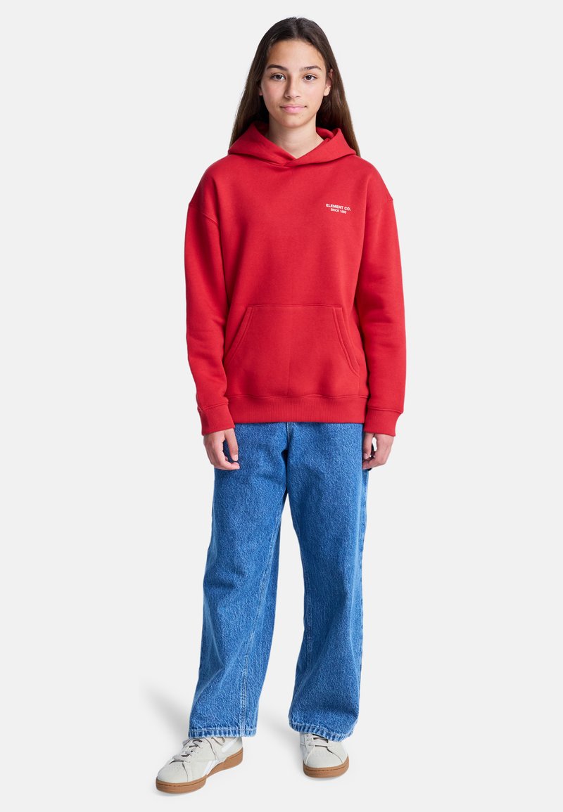 Ragazzina in piedi, che indossa una felpa rossa con cappuccio e tasca frontale, jeans blu e sneaker bianche su uno sfondo bianco semplice.