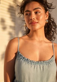 Himmelblaues, ärmelloses Kleid mit rüschigem Ausschnitt, aus weichem Stoff. Runde goldene Ohrringe ergänzen das Aussehen vor einem neutralen Hintergrund.