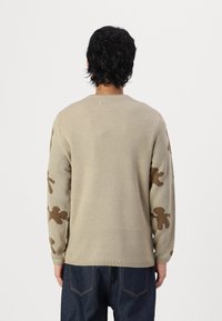 Beige knit sweater with brown bear pattern on sleeves, ribbed crew neckline, and soft texture. Casual fit, shown from the back.