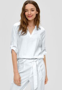 Blouse en lin blanc avec un col en V et des manches retroussées, associée à un pantalon assorti avec une ceinture nouée. Texture douce et légère.