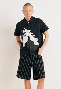 Black short-sleeve shirt with white horse graphic, displayed with black and white striped shorts. Features button closure and relaxed fit.
