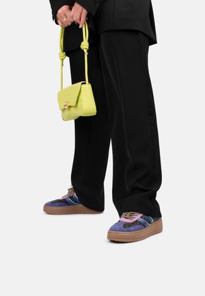 Sneaker viola in camoscio con accenti multicolori, suola beige e lacci intricati, abbinate a una piccola borsa gialla chiaro tenuta da una persona.