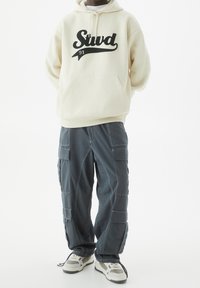 Personne portant un hoodie crème avec le texte "Stud 91", un pantalon cargo gris foncé et des baskets blanches, se tenant devant un fond blanc.