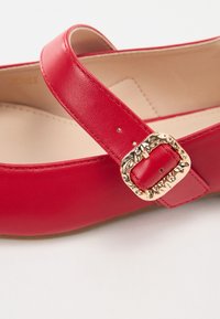 Bailarina de cuero rojo con punta redonda, que presenta una tira ancha y un detalle de hebilla en tono dorado. Textura suave con un interior beige.