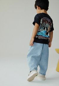 Mustat graafiset t-paidat, joissa on palmuja ja aaltoja, yhdistettynä vaaleansinisiksi löysiin farkkuihin ja valkoisiin lenkkareihin vihreillä yksityiskohdilla.