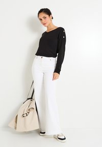 Mujer con top negro de manga larga y jeans blancos de pierna ancha, sosteniendo un abrigo beige y un bolso de leopardo, de pie contra una pared blanca.
