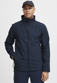 Marine blå softshell-jakke med høy krage, full glidelås foran, sidelommer og glatt tekstur. Modellen har på seg en matchende caps.