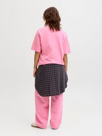 Personne aux cheveux bruns ondulés portant un t-shirt rose ample, un pantalon large rose, des baskets blanches et une chemise à carreaux grise nouée autour de la taille, debout.