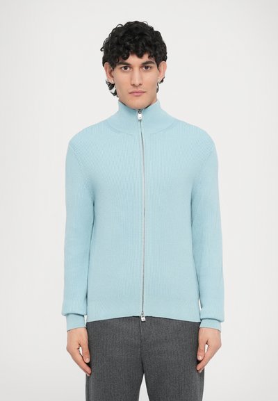 Light blue zip-up sweater in ribbed knit, featuring a high collar and long sleeves. Worn with light gray, tailored trousers.
