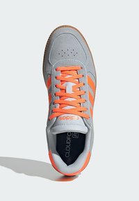 Sivá športová obuv s oranžovými akcentmi, s semišovým zvrškom, gumovou podrážkou a šnurovaním so šiestimi očkami. Povrch má textúru a je viditeľné logo.