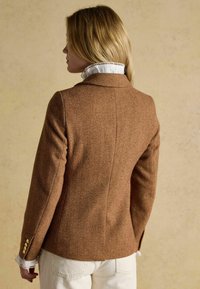 Sacou bej în model herringbone, cu o croială structurată, guler cu încrețituri și butoane decorative aurii pe mâneci. Cămașă albă cu guler vizibil dedesubt.