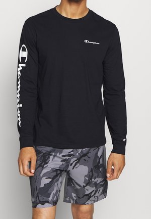 Un homme porte un t-shirt Champion noir à manches longues avec un logo sur la poitrine et la manche, assorti à un short camouflage gris et noir.