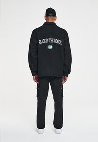Chaqueta negra con solapa, que presenta el texto blanco "PLACE IN THE WOODS" cosido en la parte posterior. También, pantalones cargo negros y zapatillas blancas.