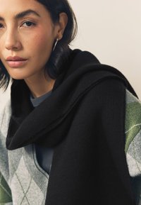 Overdimensioneret sort sjal draperet over et gråt og grønt mønstret tøjstykke. Glat stof med synlige kanter og lagdelt design.