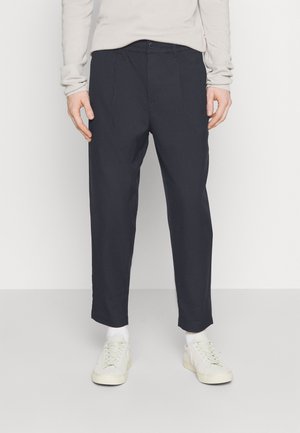 Pantaloni tapered blu navy con pieghe frontali, realizzati in tessuto leggero. Indossati con sneakers bianche e un maglione di colore chiaro.
