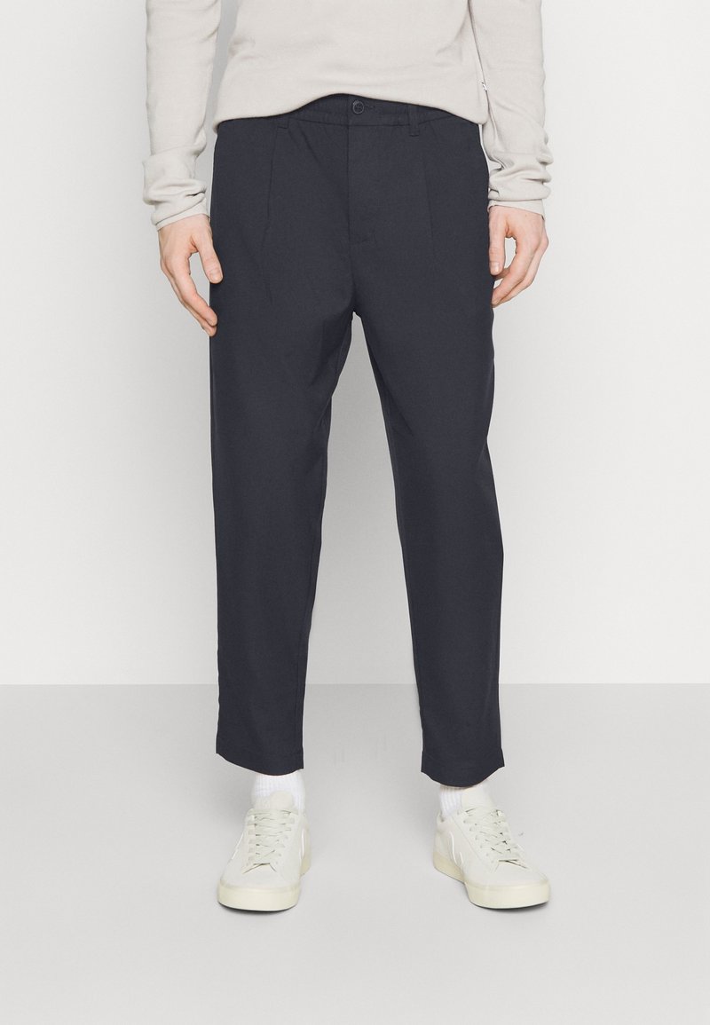 Marinblå tapered byxor med framfickor, tillverkade av lättviktsmaterial. Bärs med vita sneakers och en ljusfärgad tröja.