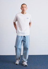 Tricou alb din bumbac cu un mic logo albastru, purtat cu blugi lejeri de culoare albastră deschisă și adidași gri, stând în fața unui fundal simplu.