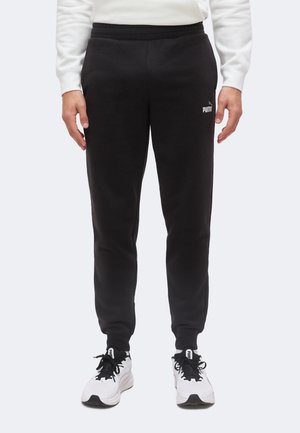 Persoană purtând pantaloni de trening negri Puma, hanorac alb și adidași albi Puma, stând în picioare cu fața spre înainte, pe un fundal simplu și deschis la culoare.