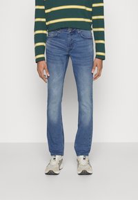 Blå denim jeans med rett passform, med en falmet vask, fem lommer og knappelukking. Brukt med en grønnstripet langermet skjorte.