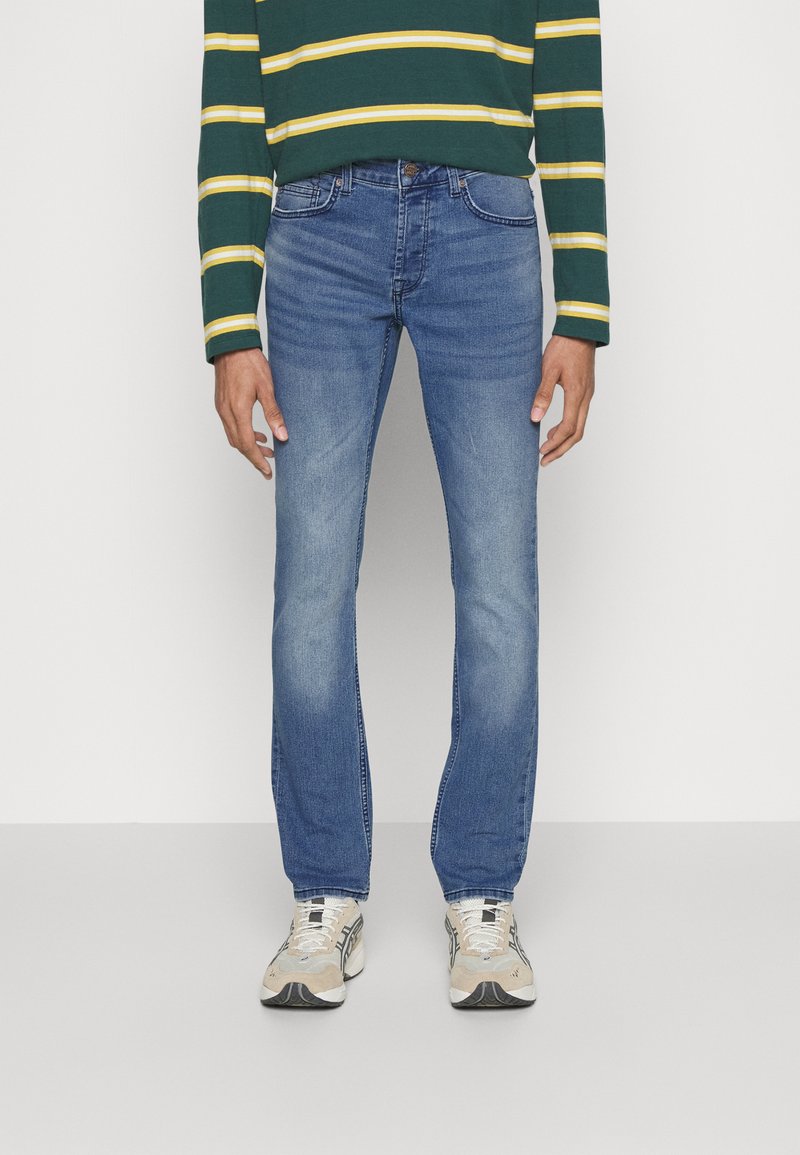 Blå denim jeans med rett passform, med en falmet vask, fem lommer og knappelukking. Brukt med en grønnstripet langermet skjorte.