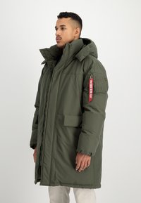 Mand iført lang, olivenfarvet polstret parka med høj krave og et rødt "Remove Before Flight" mærke på lommen på venstre ærme, stående mod en hvid baggrund.