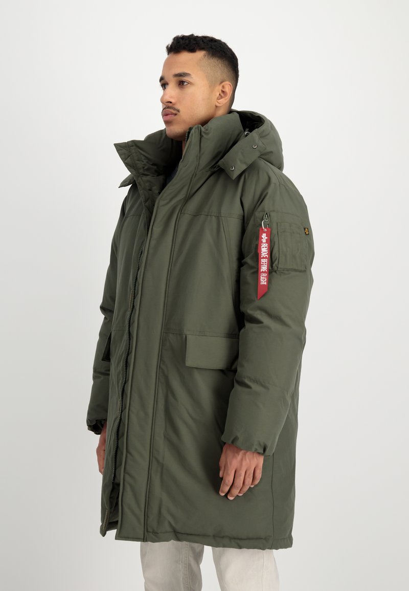 Mand iført lang, olivenfarvet polstret parka med høj krave og et rødt "Remove Before Flight" mærke på lommen på venstre ærme, stående mod en hvid baggrund.
