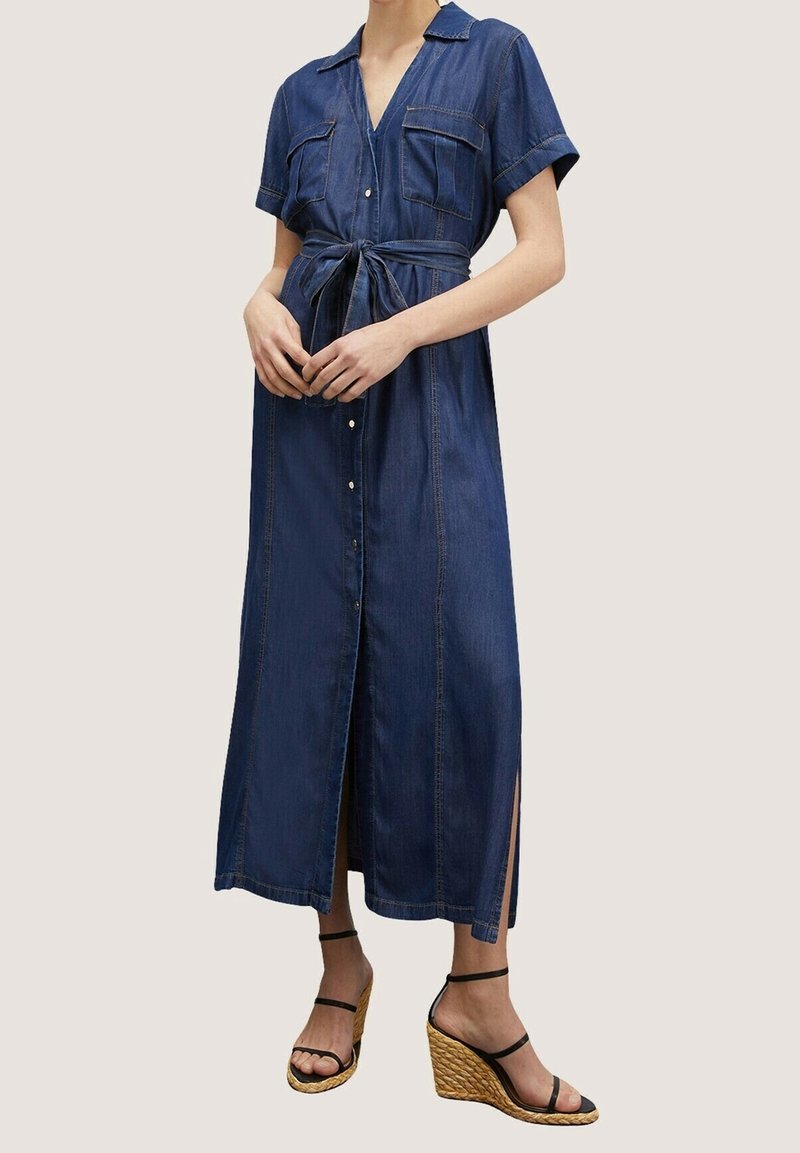 Jeanskjol i mörkblått med korta ärmar, två bröstfickor, midjeresning, knappstängning fram och sidofickor. Bärs med klacksandaler.