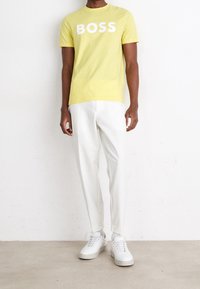 Man wearing yellow T-shirt with "BOSS" text, white pants, white socks, and white sneakers standing against a white wall and beige floor.