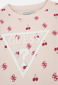 T-shirt di cotone rosa chiaro con piccoli motivi di ciliegie rosse, un logo triangolare bianco con la scritta "GUESS U.S.A." e una grafica testurizzata.