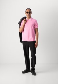 Rosa polo skjorte med krage, kombinert med svarte bukser og sko. Modellen holder en svart lærjakke og har på seg mørke solbriller.