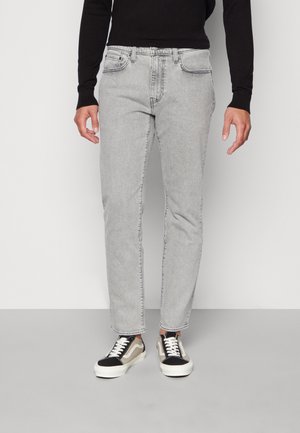 Person trägt hellgraue Jeans, schwarze und beige Sneaker und ein schwarzes Langarmshirt und steht vor einem schlichten weißen Hintergrund.