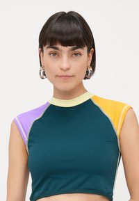 Crop top i teal med lilla, gul og oransje aksenter. Har korte ermer, høy krage og kontrasterende sømdetaljer.