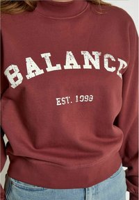 Sudadera burdeos con cuello alto; "BALANCE" y "EST. 1998" en texto blanco desgastado, hecha de una tela suave con un ajuste relajado.