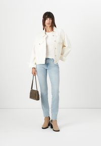 Veste en denim crème, top blanc ajusté, jean bleu clair, sac à main marron à motif zigzag et bottines à talons beiges.