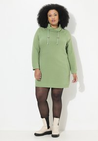 Robe à capuche verte avec col montant, manches longues, poches et cordons de serrage ajustables. Portée avec des collants noirs et des bottes à talons beiges.