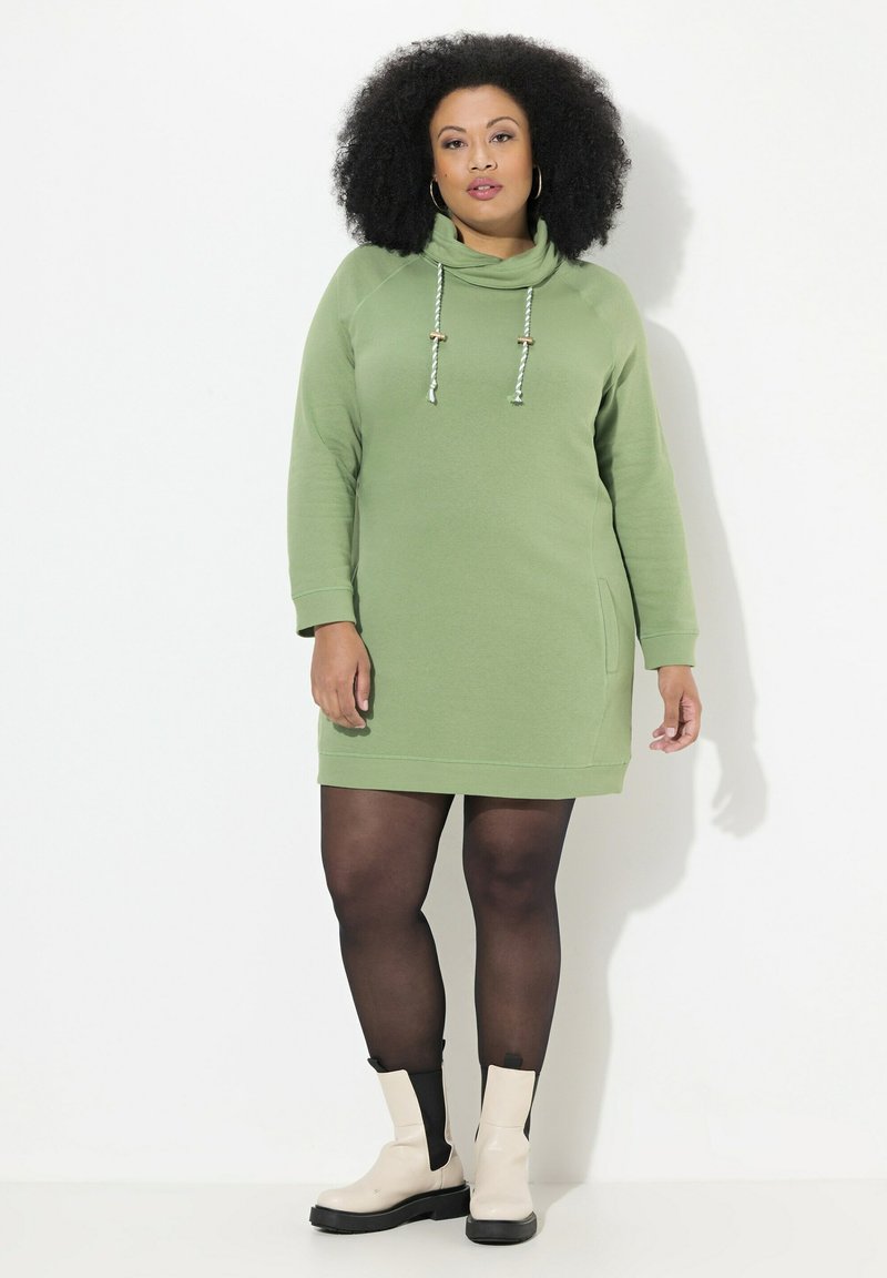 Robe à capuche verte avec col montant, manches longues, poches et cordons de serrage ajustables. Portée avec des collants noirs et des bottes à talons beiges.