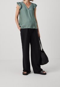 Haut vert clair texturé avec manches à volants, associé à un pantalon noir à jambes larges. Sac noir et sandales avec accents argentés complètent le look.