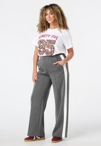 Grijze wijde broek met witte zijstrepen, gecombineerd met een wit T-shirt met een rood en panterprint nummer 95. Casual schoeisel.