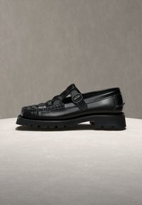 Mocassin en cuir noir avec une bride à boucle, une semelle épaisse et un détail tissé sur la partie supérieure, posé sur une surface claire avec un fond gris.