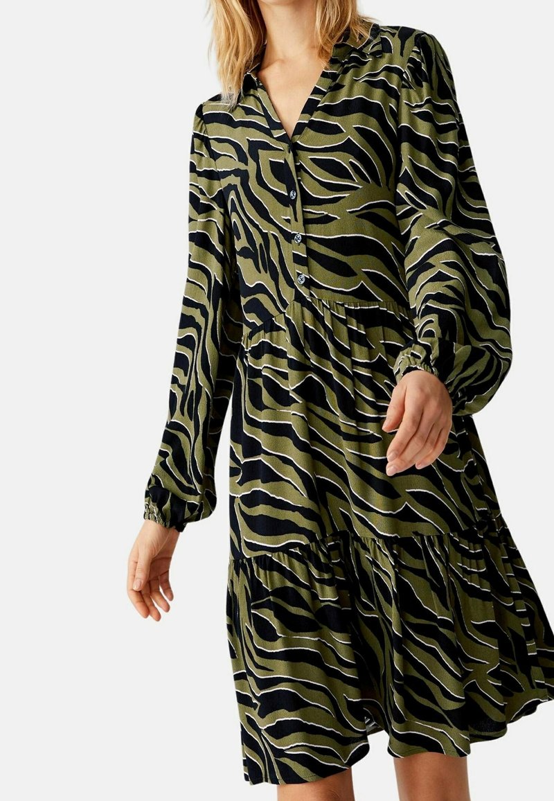 Femme portant une robe boutonnée à manches longues avec un motif zèbre vert olive et noir, jupe à volants et poignets élastiques.