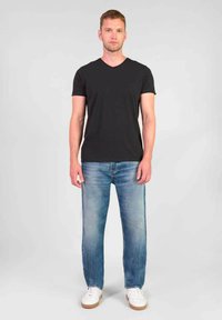 Zwarte V-hals T-shirt, lichtblauwe rechte spijkerbroek en witte sneakers met beige zolen. Man staat tegen een neutrale achtergrond.