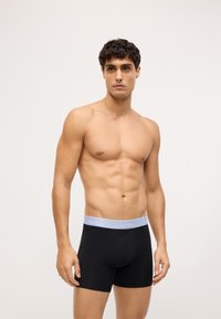 Čierne boxerky s svetlošedým elastickým pásom s logom "Calvin Klein", zobrazené na mužskom modeli na jednoduchom pozadí.