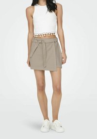 Weißes, kurzes Tanktop mit gewelltem Saum, kombiniert mit einem beigen Wickelrock mit Bindeband in der Taille, ergänzt durch weiße Turnschuhe.