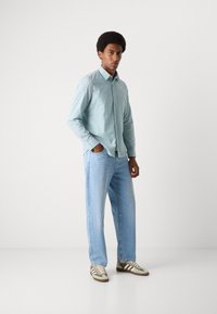 Cămașă button-up cu dungi albastre deschise, blugi albaștri deschise și adidași bej cu accente verzi și maronii, purtați de o persoană în picioare.