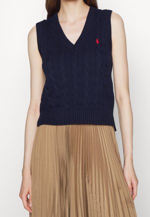 Kobieta ubrana w granatową kamizelkę na szydełkowym wzorze bez rękawów z dekoltem w serek i beżową plisowaną spódnicę, widoczna od ramion do bioder.