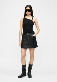 Donna che indossa un top nero asimmetrico senza maniche, gonna di pelle nera con bottoni sul davanti, stivali neri fino a metà polpaccio e occhiali da sole neri oversize.