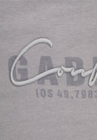 Šedá tkanina s vyšitým textem a stylizovaným logem. Textura je hladká, s vystouplým stehem v světlejším odstínu.