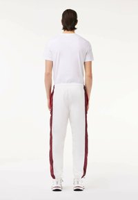 Vyras stovi atsigręžęs, vilkintis baltą marškinėlį, baltas sportines kelnes su raudonomis šoninėmis juostelėmis ir baltus sportinius batelius ant vienspalvio fono.