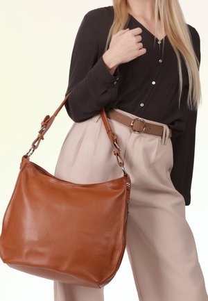 Woman wearing black blouse and beige trousers holding large brown leather shoulder bag with metal hardware.
